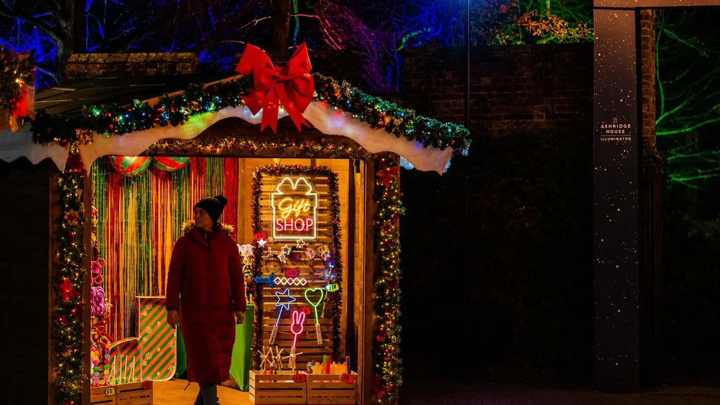 A visitor outside a gift shop hut decked with garland, a red bow and a neon Gift Shop sign, on the Ashridge House Illuminated trail